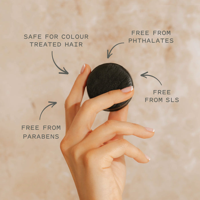 A hand holds the Good Juju Body & Home Scalp Care Conditioner Bar—a black, round bar with text: Safe for colour treated hair, Free from phthalates, SLS & parabens; a natural conditioner for gentle scalp care.