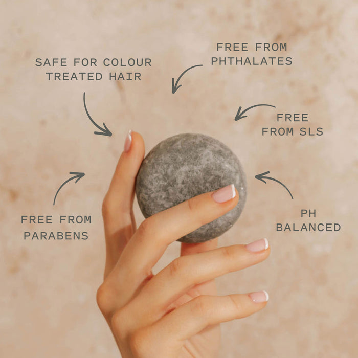 A hand with manicured nails holds a round gray Scalp Care Conditioner Bar by Good Juju Body & Home. Text: Safe for colour treated hair, pH balanced, free from parabens, phthalates & SLS; relieves dandruff and pairs with our natural shampoo bar.