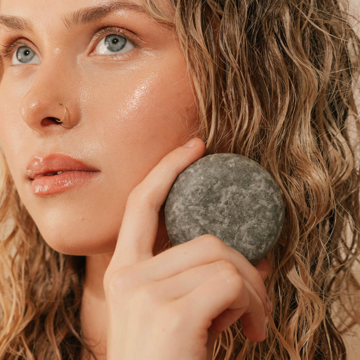 A woman with wet, wavy blonde hair holds a gray stone to her cheek. Her fresh look is enhanced by Good Juju Body & Home's Scalp Care Conditioner Bar, ideal for scalp care and helping relieve dandruff.