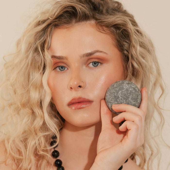 A woman with curly blonde hair holds the Good Juju Body & Home Scalp Care Conditioner Bar to her cheek. She wears light makeup and a black beaded necklace, posing against a plain, light background.