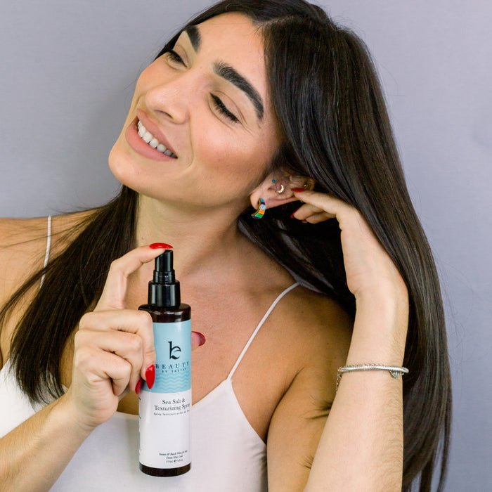 A woman with long dark hair smiles while holding a bottle of Beauty by Earths Sea Salt Spray Hair Texturizer near her hair. She wears a white tank top, colorful earrings, and a bracelet against a gray background that complements the products natural vibe.