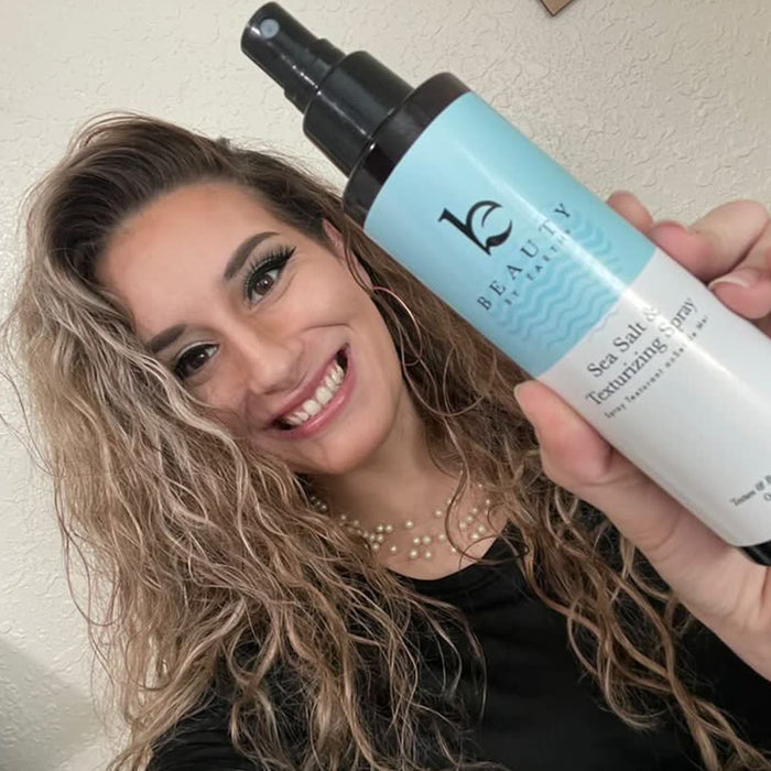 A curly-haired person holding a bottle of Beauty by Earth Sea Salt Spray Hair Texturizer smiles while wearing a black top and pearl necklace, posing against a light-colored wall.
