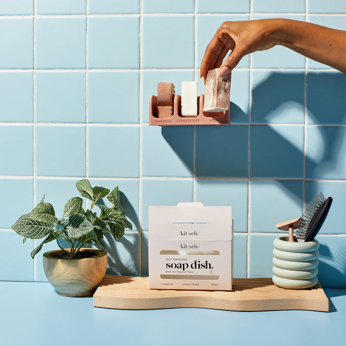 A hand places solid shampoo and conditioner bars on a pink holder against a light blue tiled wall, highlighting bottle-free beauty. Below is the Kitsch Self Draining Soap Dish, a small plant pot, and a toothbrush holder with brushes on the wooden surface.