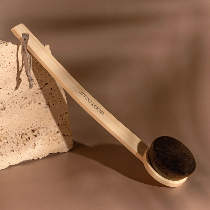 An EcoTools Self-Tan Back Applicator with a long wooden handle and dark bristles is on a brown surface next to a textured stone. It has a fabric loop and features ecotools, perfect for applying your favorite self-tanner.