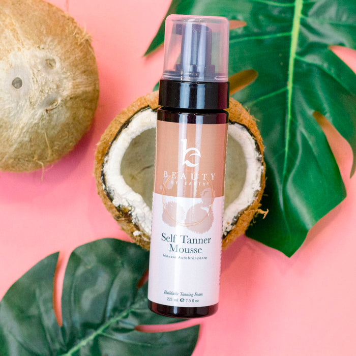 A Beauty by Earth Self Tanner Mousse bottle is placed before a coconut half on a pink backdrop, surrounded by green leaves for a tropical feel. The label assures natural ingredients for a desired sun-kissed glow.