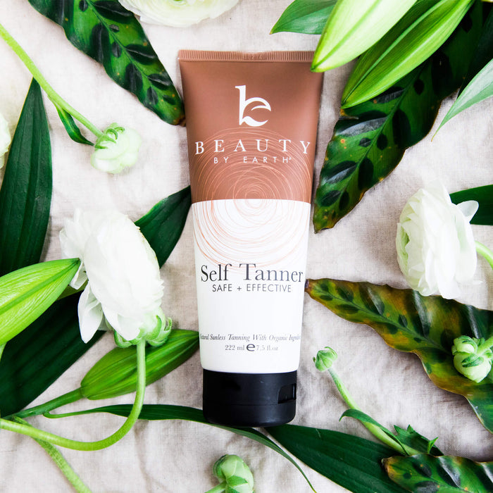 A Beauty by Earth Self Tanner tube rests on textured fabric, encircled by green leaves and white flowers. The brown and white packaging ensures a safe, effective sunless tanning method for a golden glow.