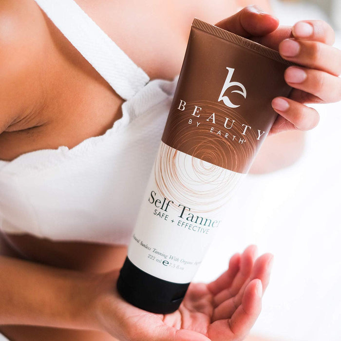 Elegantly posed in a white top, a person holds Beauty by Earths Self Tanner, showcasing its promise for a safe and effective sunless tan. The brown and white tube hints at the golden glow within, graced by their poised hand.