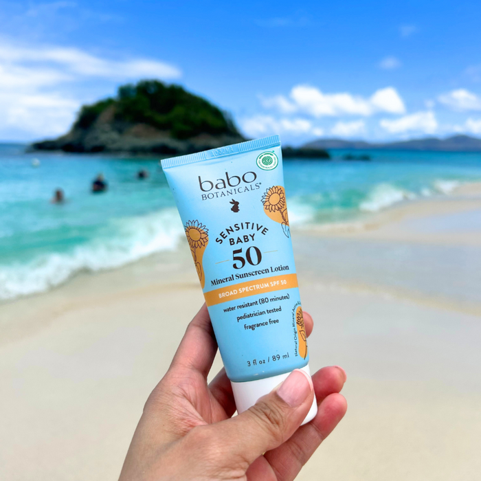 A hand holds Babo Botanicals Sensitive Baby Mineral Sunscreen Lotion SPF50 on a sandy beach, with turquoise water and a lush island in the background under a blue sky.