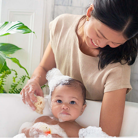 A woman smiles while giving a bubble bath to a baby in a white tub using Babo Botanicals Sensitive Baby Shampoo & Wash. The baby is surrounded by bubbles with soapy foam on their head, set against a lush backdrop of green plants, ensuring the moment is gentle on sensitive skin.