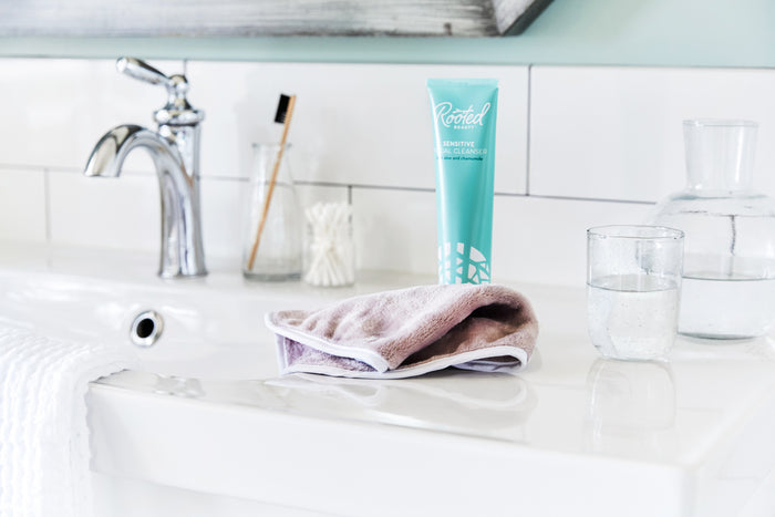 A bathroom sink with a silver faucet features a tan washcloth, toothbrush in a holder, cotton swabs, and two water-filled clear glasses on the countertop. A teal tube labeled Sensitive Skin Facial Cleanser from Rooted Beauty boasts R7 complex and comes in recyclable packaging.