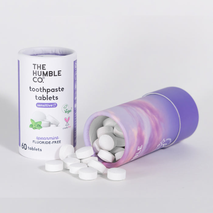 Two cylindrical containers of The Humble Co. Sensitive Toothpaste Tabs, labeled sensitive and spearmint, are displayed. One container stands upright while the other is tipped over, revealing eco-friendly packaging tablets. Both are white and purple with 60 tablets each.