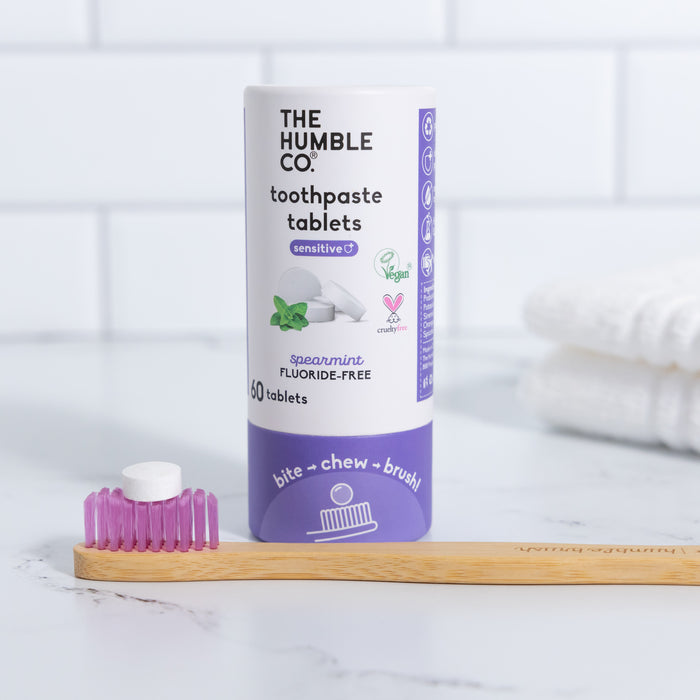 A cylindrical container of The Humble Co. Sensitive Toothpaste Tabs, fluoride-free and in eco-friendly packaging labeled vegan and cruelty-free, is on a white bathroom countertop. A bamboo toothbrush with pink bristles rests beside it, with white towels in the background.