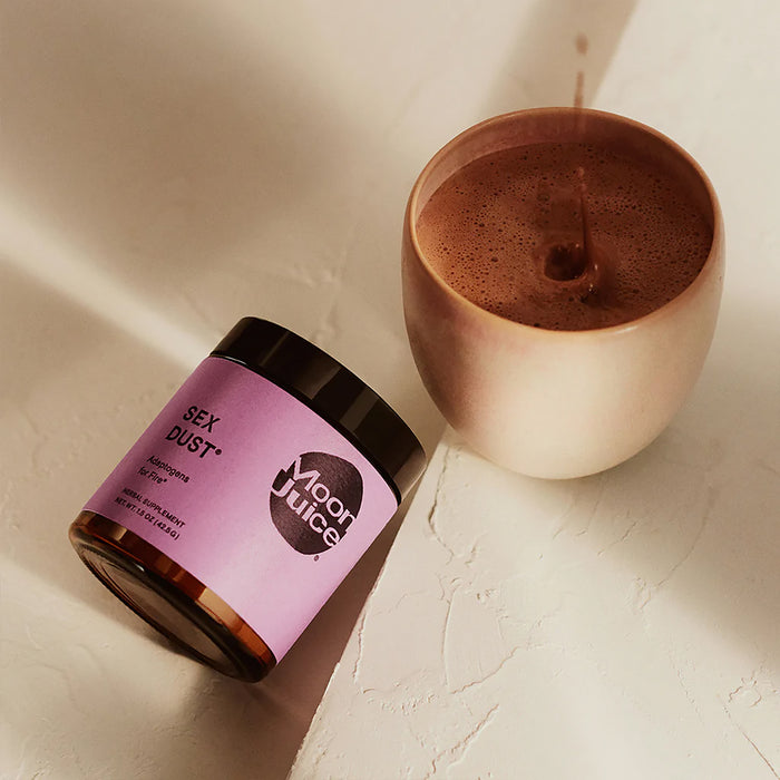 A container of Sex Dust - Adaptogens for Libido, Hormonal Balance & Creative Energy by Moon Juice, with a purple label and black lid, sits on a textured surface near a cup of frothy chocolate-colored liquid.