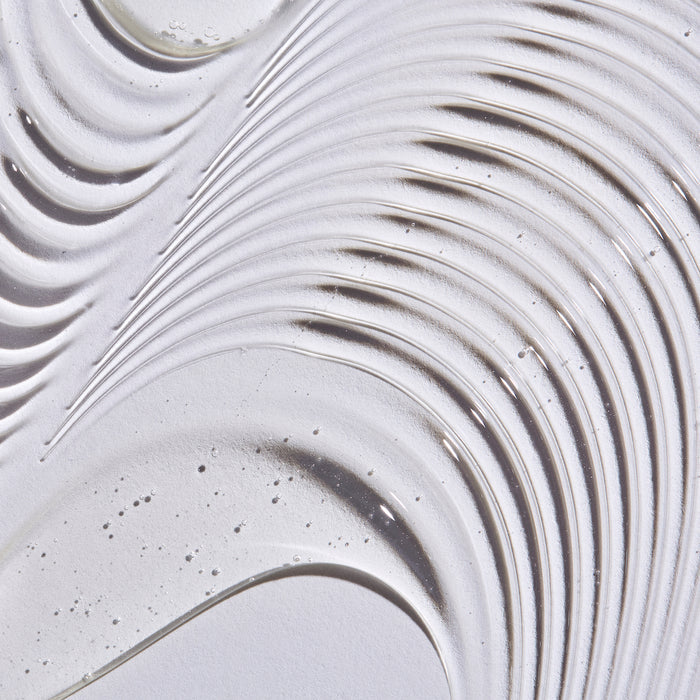 A close-up of smooth, layered swirls and curves on a textured white surface, resembling sand ridges or icing. Infused with Aloe + Chamomile Extract, bubbles and specks add depth to the abstract pattern. This resembles The Honest Companys Shampoo + Body Wash.
