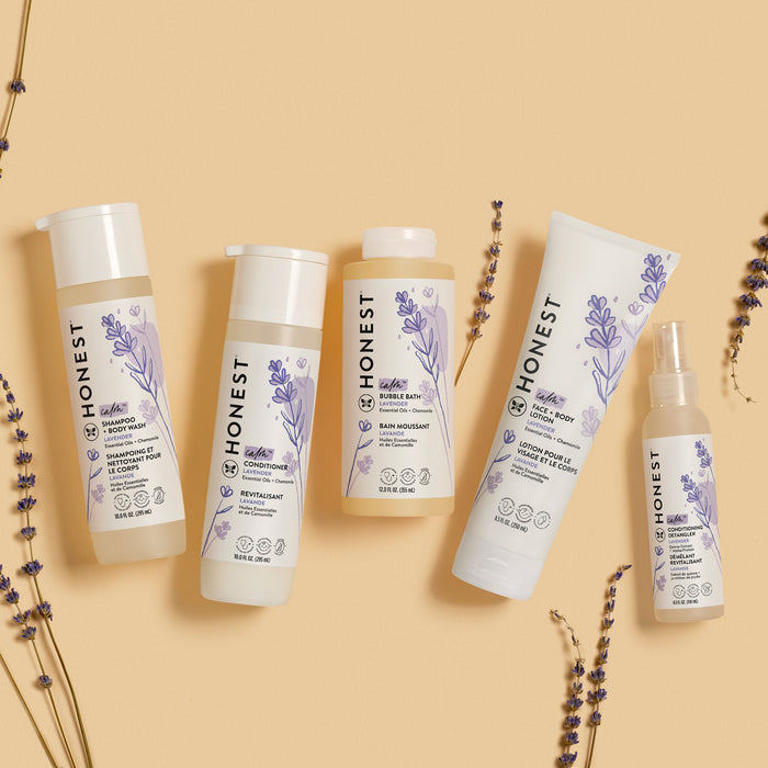 On a beige background, five bottles from The Honest Company showcase lavender graphics: Shampoo + Body Wash, conditioner, bubble bath, face and body lotion, and conditioning detangler. Lavender sprigs and hints of Aloe + Chamomile Extract are scattered around.