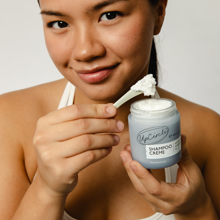 A smiling person in a white top holds UpCircles Shampoo Crème with Coconut + Grapefruit Oil in one hand and a small spatula of the product in the other, embodying the Shampoo Revolution against a white background.