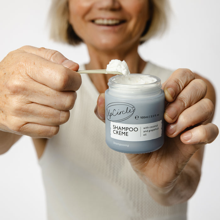 A person in a light sleeveless top holds a jar of UpCircles Shampoo Crème with Coconut + Grapefruit Oil, representing the Shampoo Revolution. Using a small spatula, they scoop out some of the hybrid crème from the jar labeled with natural ingredients like coconut and grapefruit oil.