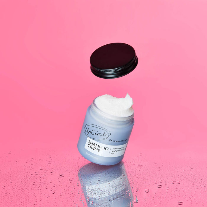 A jar of UpCircles Shampoo Crème with Coconut + Grapefruit Oil, featuring a black lid, hovers open against a pink background. White cream seems to float out of it, with a reflective surface dotted by water droplets, symbolizing a natural ingredients revolution.