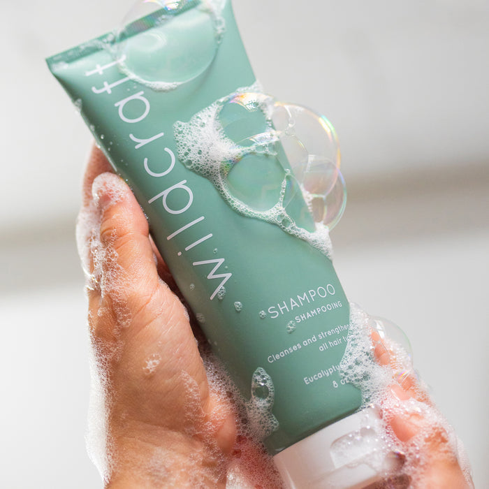 A hand holds a green tube of Wildcraft Shampoo with eucalyptus and mint, covered in soap bubbles. The label states, Cleanses and strengthens all hair types with quinoa protein. The background is blurred to emphasize the product.