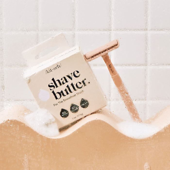 A box of Kitsch Shave Butter and a rose gold safety razor sit on a foamy bathtub ledge against a minimalist, bottle-free bathroom with white tiled walls.