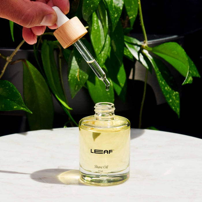 A hand uses a dropper to release a moisturizing drop of yellow Shave Oil w/ Dropper by Leaf Shave into a glass bottle on a white surface, with softly focused green leaves in the background.