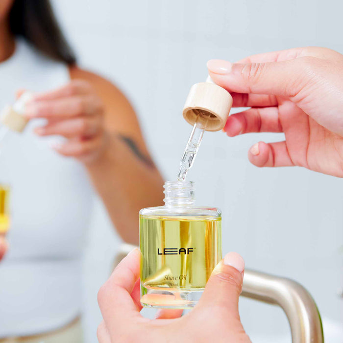 Close-up of someone holding a dropper above a Leaf Shave Shave Oil w/ Dropper bottle, showcasing its irritation-reducing and moisture-locking qualities. In the background, another person holds a similar bottle in an ideal bathroom self-care setting.
