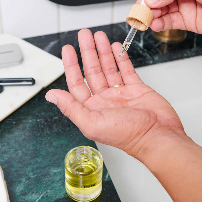 Against a green marble backdrop, a person uses the dropper from Leaf Shaves Shave Oil w/ Dropper to apply moisture-rich oil onto their open palm.