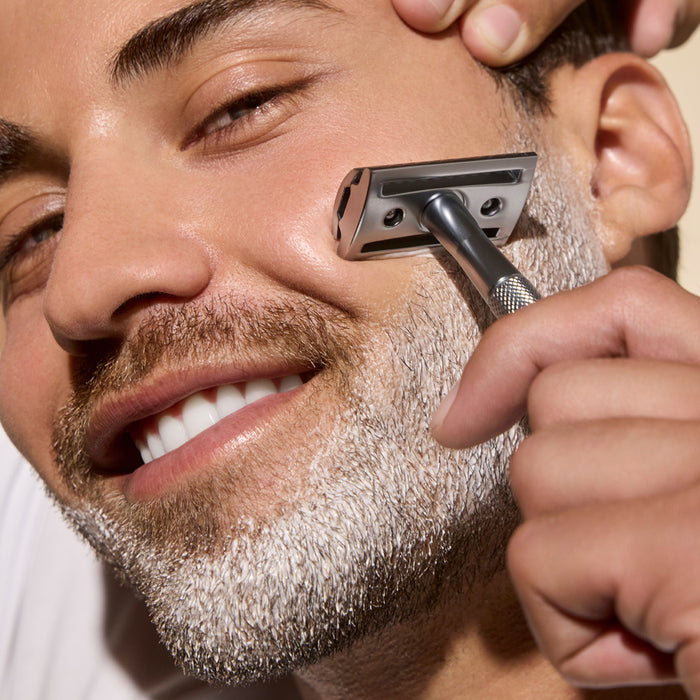 A smiling man is shaving his beard with a metal razor using Phil's Shaving Cream, while another hand gently touches his forehead.