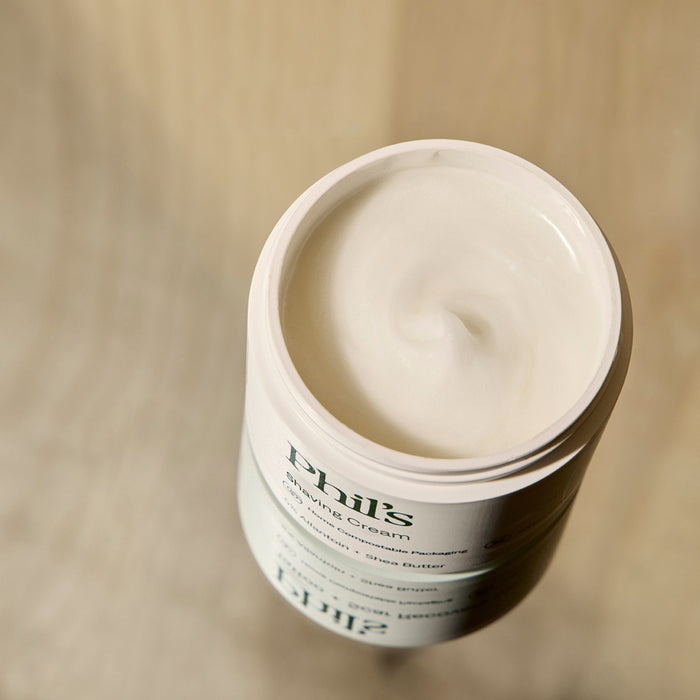 An open container of Phil's Shaving Cream rests on a beige surface, displaying the smooth, white cream inside. The label, partially visible, shows the Phil's brand and product type.