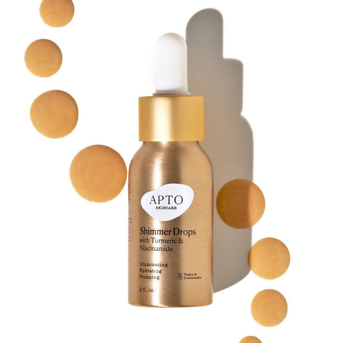A gold bottle labeled Shimmer Drops with Turmeric & Niacinamide, Glow Serum by APTO Skincare shines among shimmering golden droplets. With a white dropper cap, this cruelty-free products shadow on the white background highlights its natural ingredients.