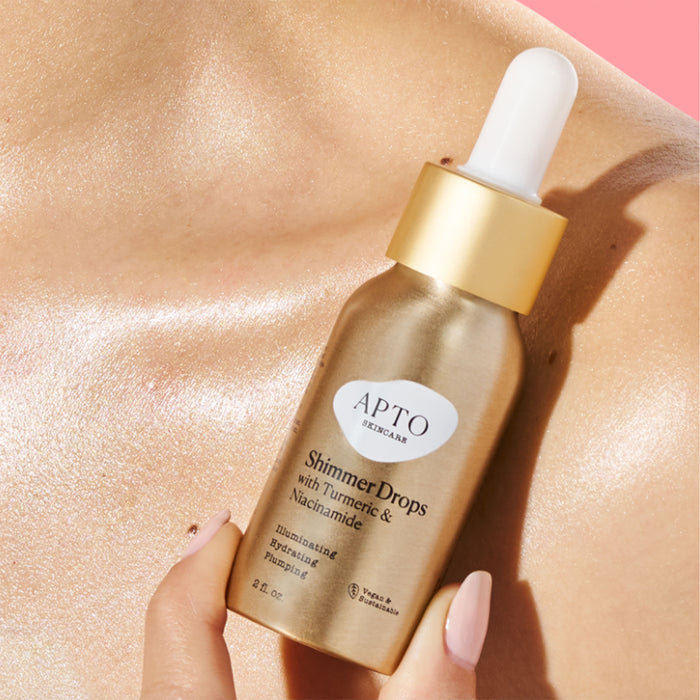 A gold bottle of APTO Skincares Glow Serum, Shimmer Drops with Turmeric & Niacinamide, featuring a white dropper cap, sits on glowing skin against a light pink background, emphasizing its natural, cruelty-free essence.