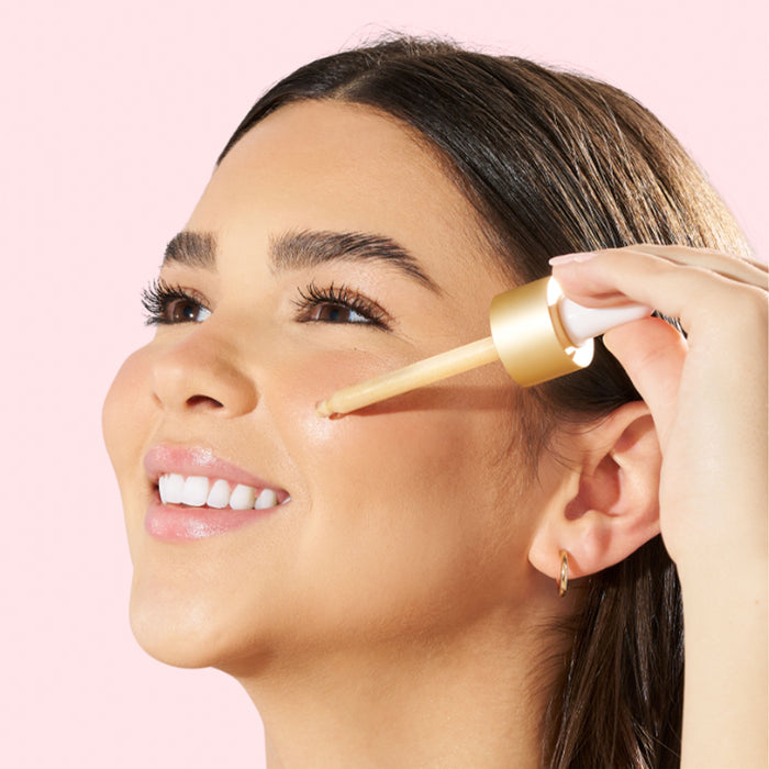 A woman with long dark hair smiles as she uses a dropper to apply APTO Skincares Shimmer Drops with Turmeric & Niacinamide, Glow Serum to her cheek. Her skin looks smooth and radiant due to its natural ingredients. The soft pale pink background enhances the scenes gentle allure.