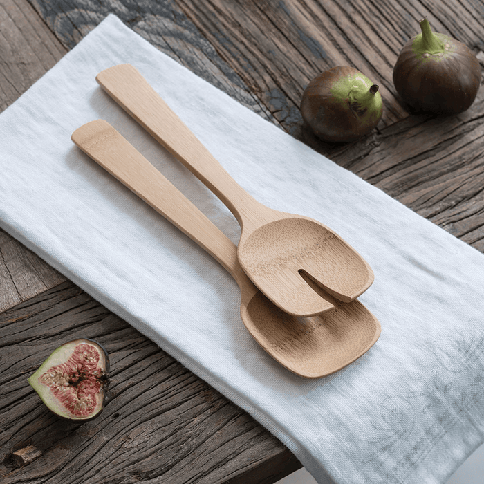 Bambus Short Servers lie on a white cloth on a rustic wooden table. Nearby, two whole figs and one halved fig complement the natural, minimalist kitchen decor, adding a hint of contemporary design.