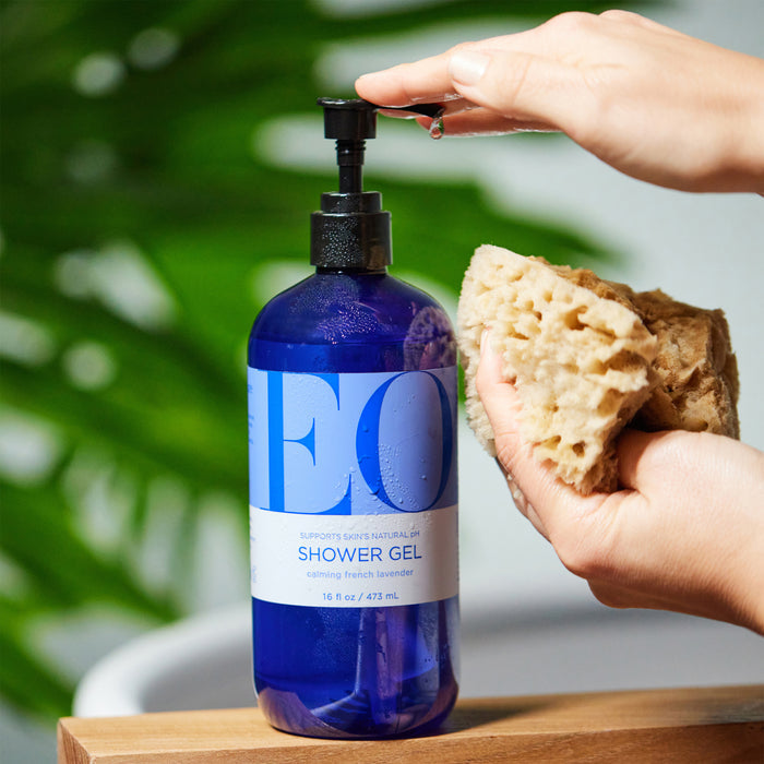 Someone uses EO Products Shower Gel, a soothing blend of Calming French Lavender in a blue bottle, on a sponge. The bottle sits on wood with a blurred green plant behind it.