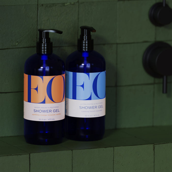 Two blue EO Products Shower Gel bottles, with pump dispensers, sit on a green tiled shelf. The left one, labeled in orange with EO in blue letters, is Jasmine Verbena. The right bottle features a blue label with EO in orange letters.