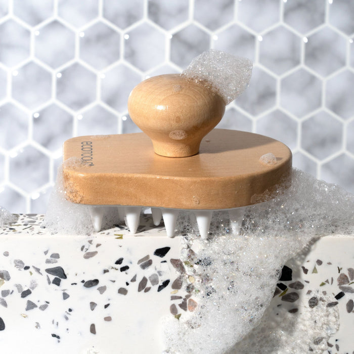 The EcoTools Shower Scalp Massager, featuring white plastic bristles for deep cleansing and hair growth, rests on a terrazzo surface with a soapy hexagonal tile background.