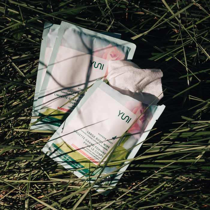 Two YUNI Shower Sheets in teal packaging, one open with a wipe partially out, rest on green grass. With a pink floral design, these sustainable sheets are perfect for on-the-go cleansing.