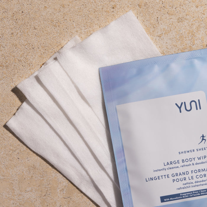 A pack of YUNI Shower Sheets sits on a beige-textured surface, with four unfolded white wipes beside it, showcasing their size and texture. Ideal for on-the-go cleansing, the packaging highlights their refreshing and deodorizing benefits with a hint of peppermint essential oil.