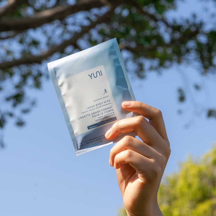 A hand holds up a YUNI Shower Sheets package, set against a backdrop of blue sky and tree branches. The body wipes, ideal for on-the-go cleansing, are white with blue and gray accents, featuring text and a small icon of a running figure.