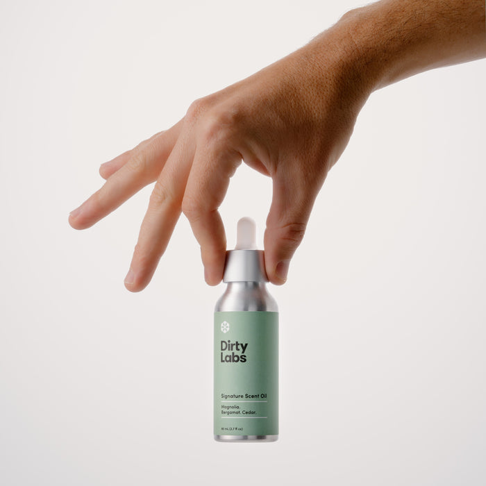 A hand delicately holds a silver bottle labeled Dirty Labs Signature Scent Oil + Glass Dropper. The elegant green label highlights premium ingredients, promising an irritant-free fragrance against a plain white background.