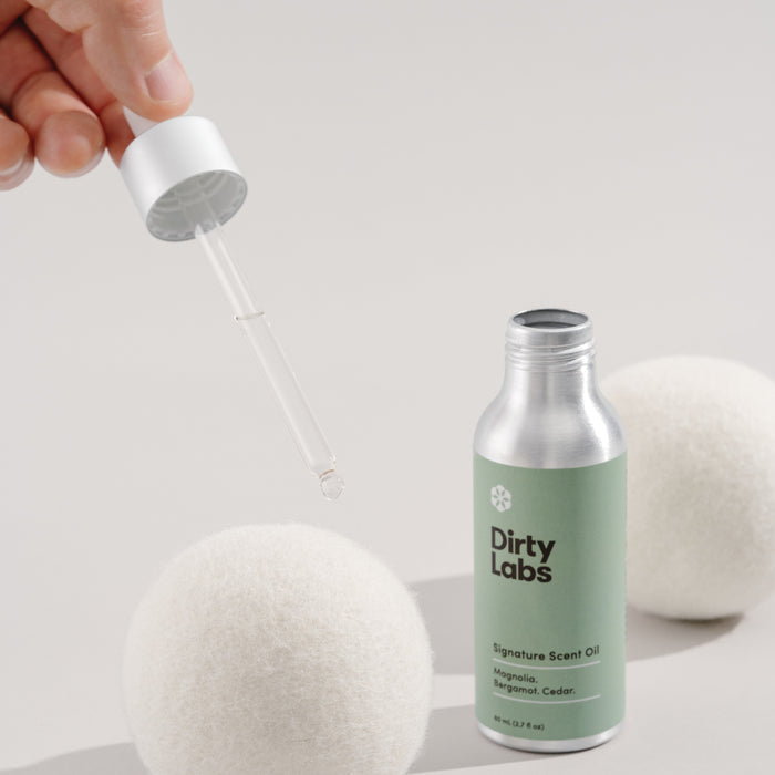 A hand holds a dropper over a wool dryer ball next to Dirty Labs Signature Scent Oil bottle. Crafted with premium ingredients, this scent offers magnolia, bergamot, and cedar aromas while being free of irritating fragrances.
