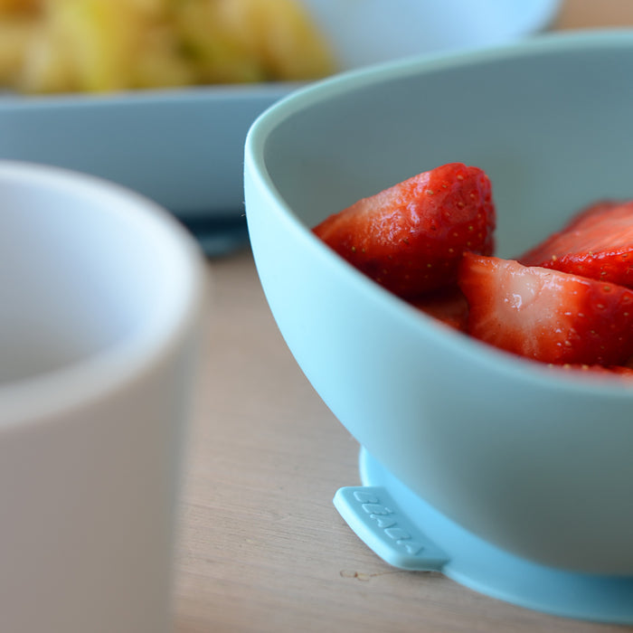 A close-up of a blue bowl filled with sliced strawberries from the BPA-free BEABA Silicone Baby Feeding Set is on a table. In the foreground, a white cup is partially visible, while another blurred bowl appears in the background, making toddler feeding safe and delightful.