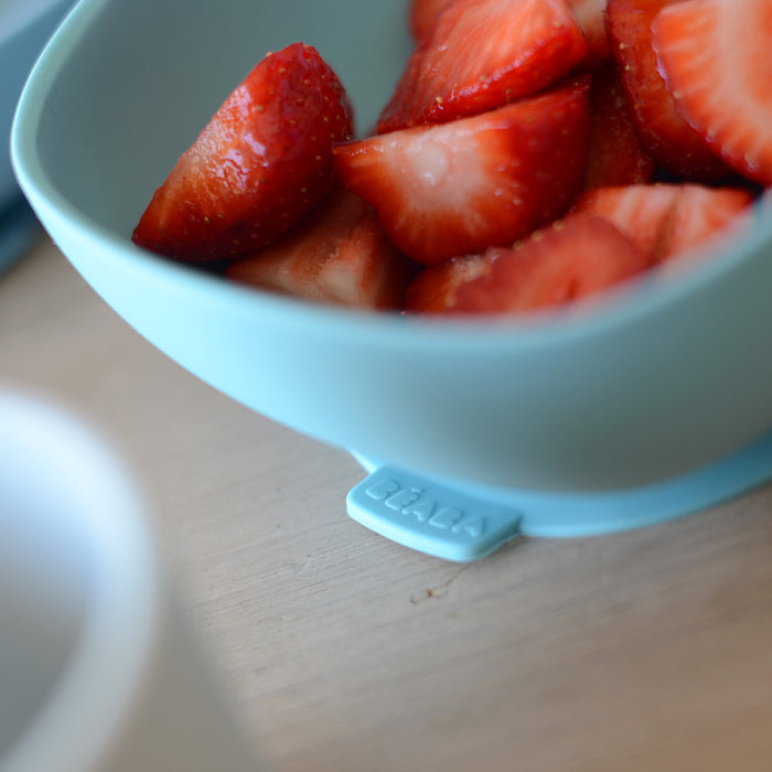 Close-up of a light blue bowl from the BEABA Silicone Baby Feeding Set, filled with sliced strawberries, showcasing their juicy texture. A blurred white object is in the foreground, perfect for toddler feeding moments. The set is BPA-free and includes a silicone suction feature.