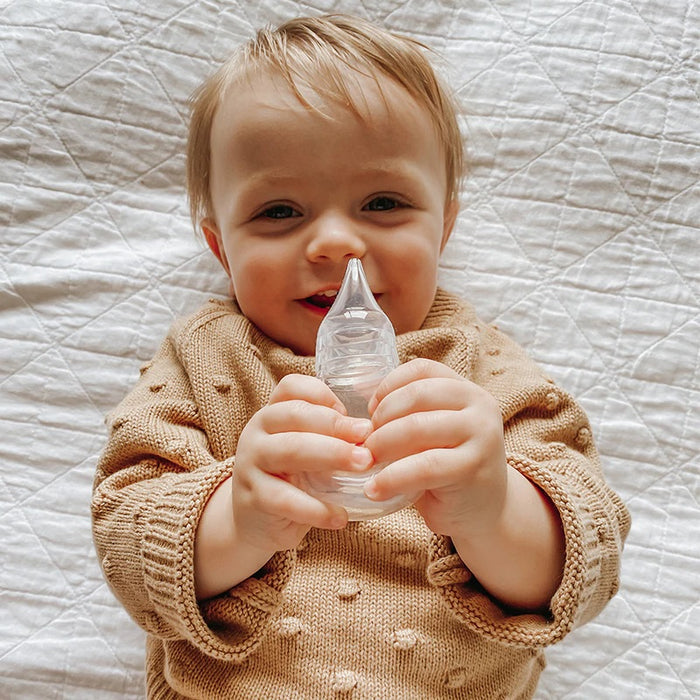 A smiling baby wearing a textured tan sweater holds a Haakaa Silicone Baby Nasal Aspirator while lying on a white quilted surface.