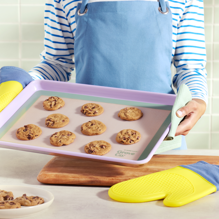 A person in a striped shirt and blue apron holds a baking tray with chocolate chip cookies on a Grove Co. eco-friendly, non-stick silicone baking mat. Nearby, yellow oven mitts rest on the counter, and more cookies sit in a bowl on a wooden board against a tiled wall backdrop.