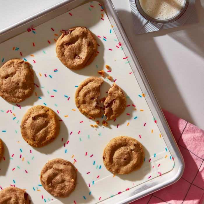 Eight chocolate chip cookies rest on a GIR Silicone Baking Mat atop a white surface with colorful decorations. One cookie is broken, and nearby, a glass of milk sits on a coaster beside an eco-friendly pink napkin featuring a geometric pattern.