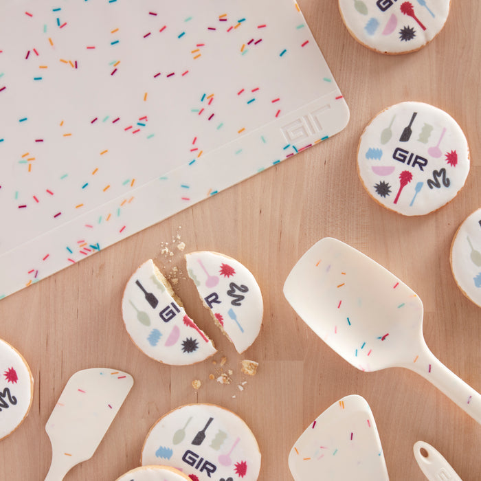 Cookies with kitchen-themed designs and colorful sprinkles rest on a GIR Silicone Baking Mat. A spatula and other eco-friendly tools, matching the sprinkle theme, are artfully arranged beside them on the wooden surface.