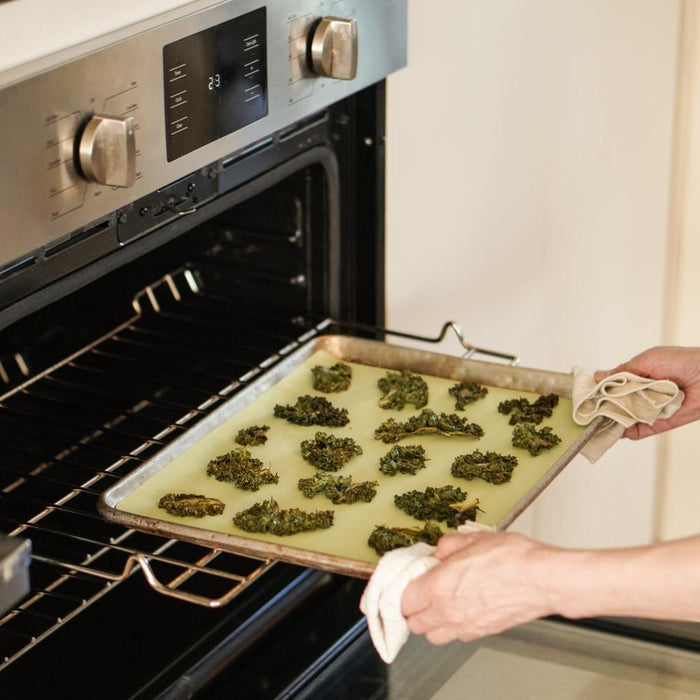 With the oven door open, someone places a tray of kale chips onto a Net Zero Co. Silicone Baking Mat, handling it with a cloth. The display shows temperature and timer settings.
