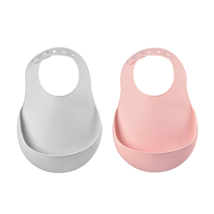 Two BEABA Silicone Bibs, essential for feeding, are shown on a white background. One is gray, the other pink; both have adjustable neck fasteners and wide food-catching pockets. Theyre also dishwasher safe for easy cleaning.