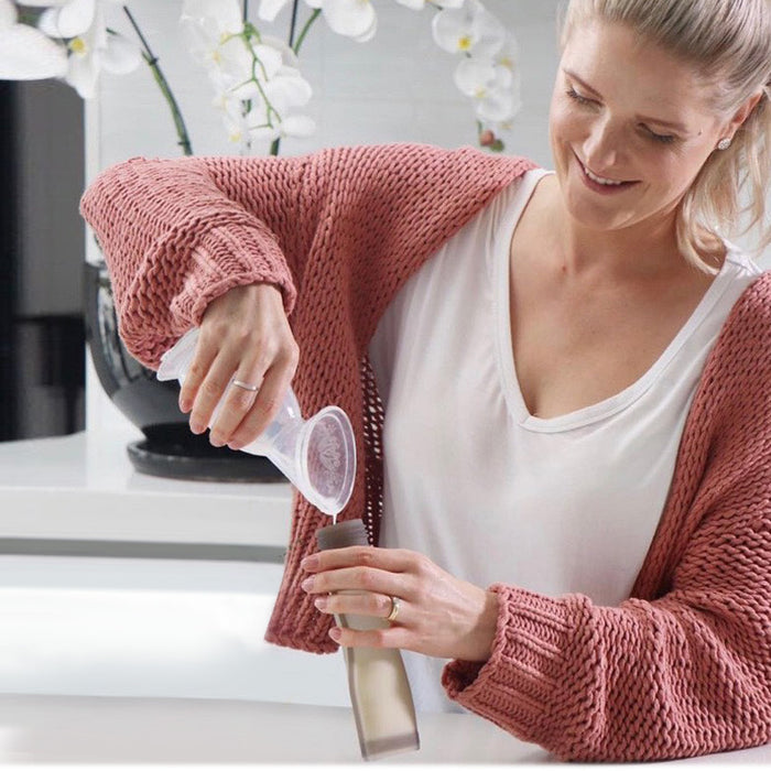 In a modern kitchen, someone in a pink sweater happily uses a funnel to pour liquid into a glass bottle—a must for eco-conscious parents. The scene is complete with Haakaas Silicone Milk Storage Bag & Sippy Spout Combo Pack and white flowers in a dark vase.
